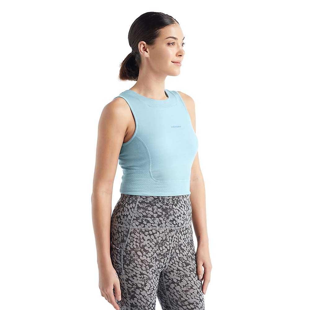 A woman with dark hair tied back wears the Icebreaker Women's ZoneKnit Cropped Bra-Tank in light blue and patterned gray breathable leggings, standing slightly to the side against a plain white background.
