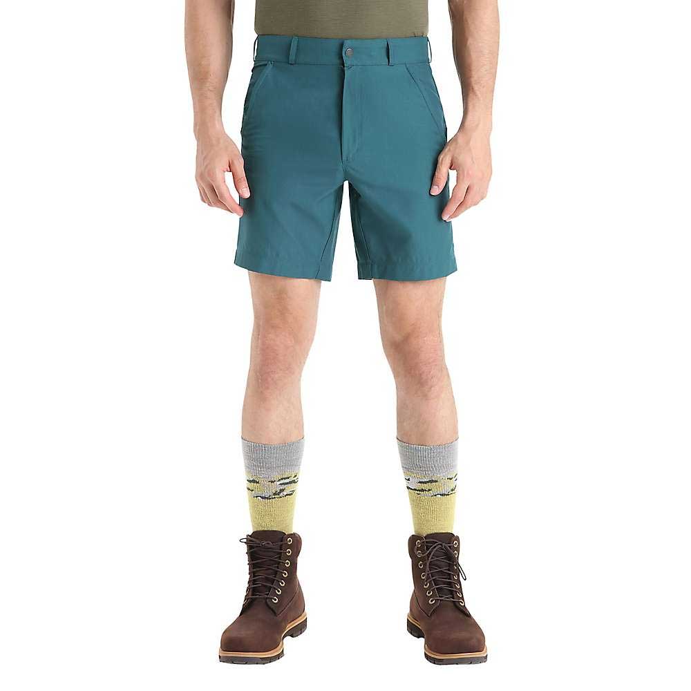 A person wearing Icebreaker Mens Hike Short in teal, an olive-green shirt, yellow and gray patterned socks, and brown lace-up boots stands with hands in pockets against a plain white background.