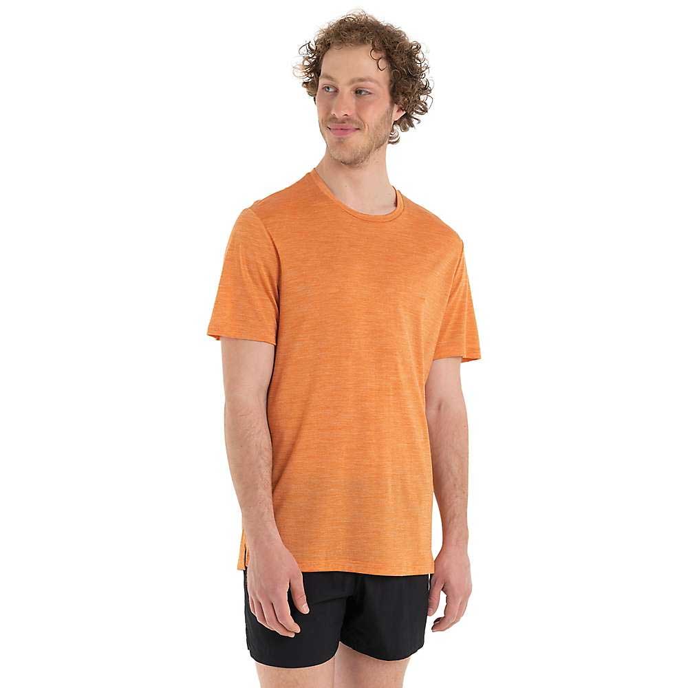 A man with curly hair, wearing the Icebreaker Men's Sphere II SS Tee in moisture-wicking orange and black shorts, stands smiling and turned slightly left against a plain white background.