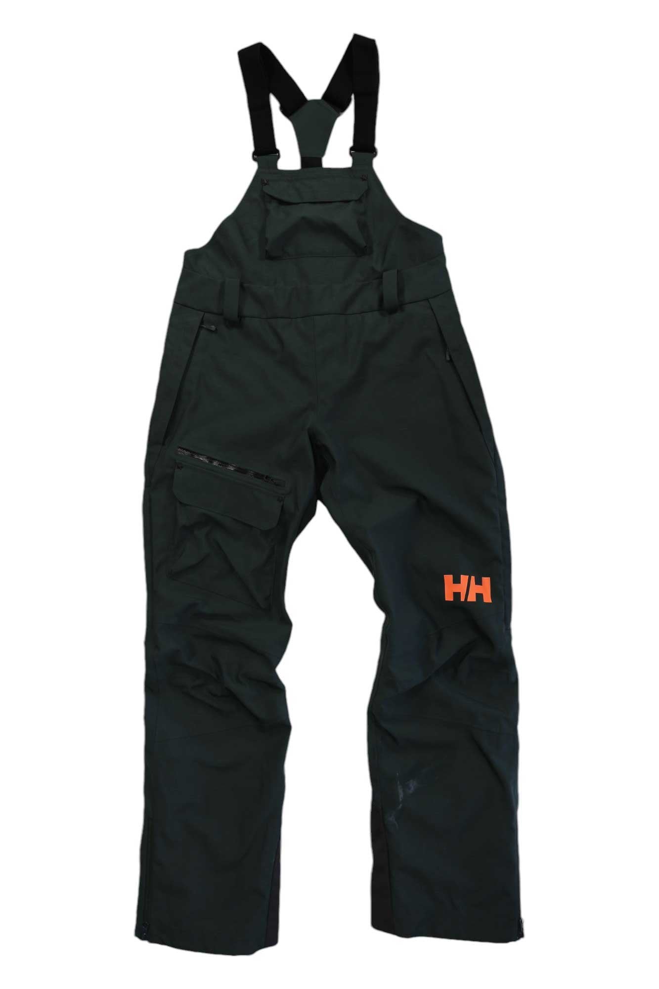 The Helly Hansen Womens Powderqueen Bib Pant in dark green has black adjustable shoulder straps, multiple pockets, an orange HH logo on the left thigh, and is made from durable, waterproof breathable 2-layer fabric for outdoor or work use.