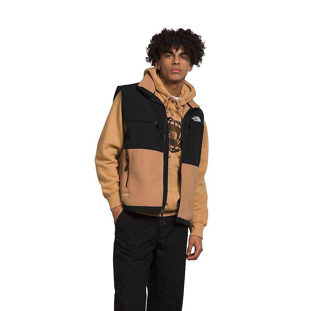 A young man with curly hair wears The North Face Men's Denali Vest in black and tan over a tan hoodie and black pants, standing with one hand in his pocket against a white background.
