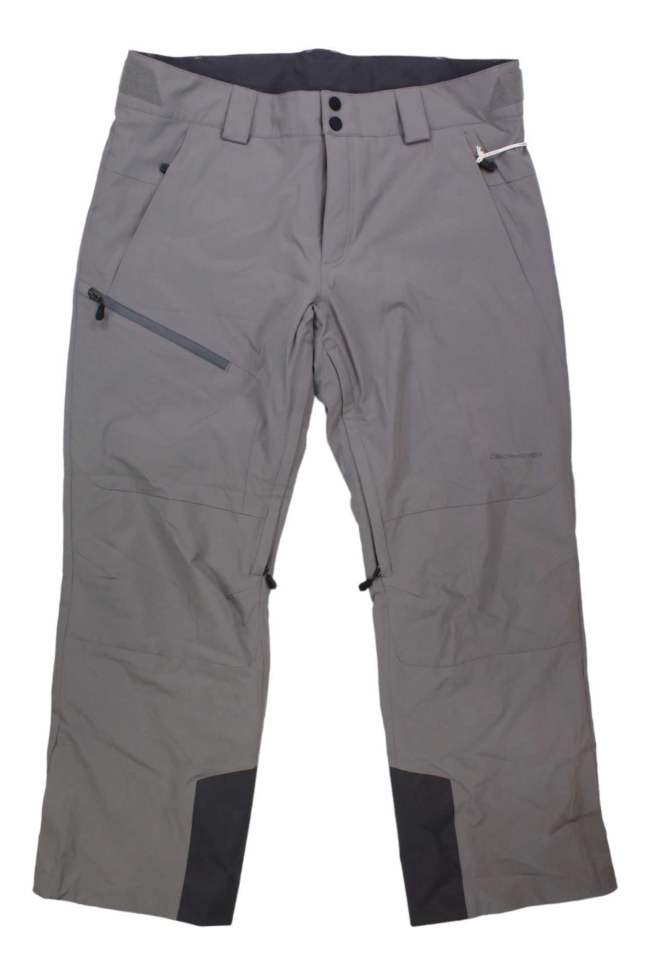 Obermeyer Men's Force Pant in gray, made with stretch fabric and HydroBlock Pro, features black reinforced inner ankle patches, front zipper pockets, belt loops, and two snap buttons at the waistband. Shown laid flat on a white background.