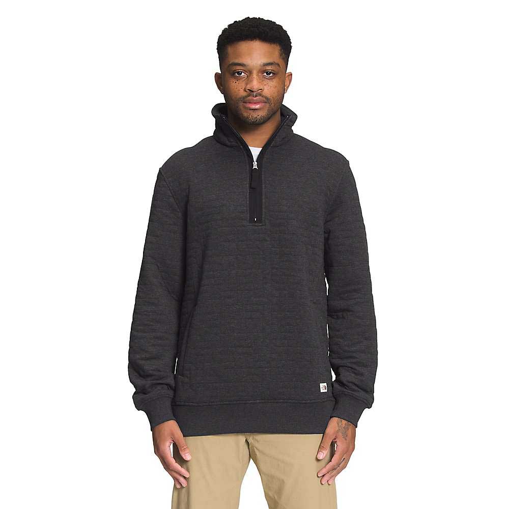 A man with short hair stands facing forward against a plain white background. He is wearing The North Face Mens Long Peak Quilted 1/2 Zip Jacket in dark gray and light tan pants. The jacket is made from recycled polyester.