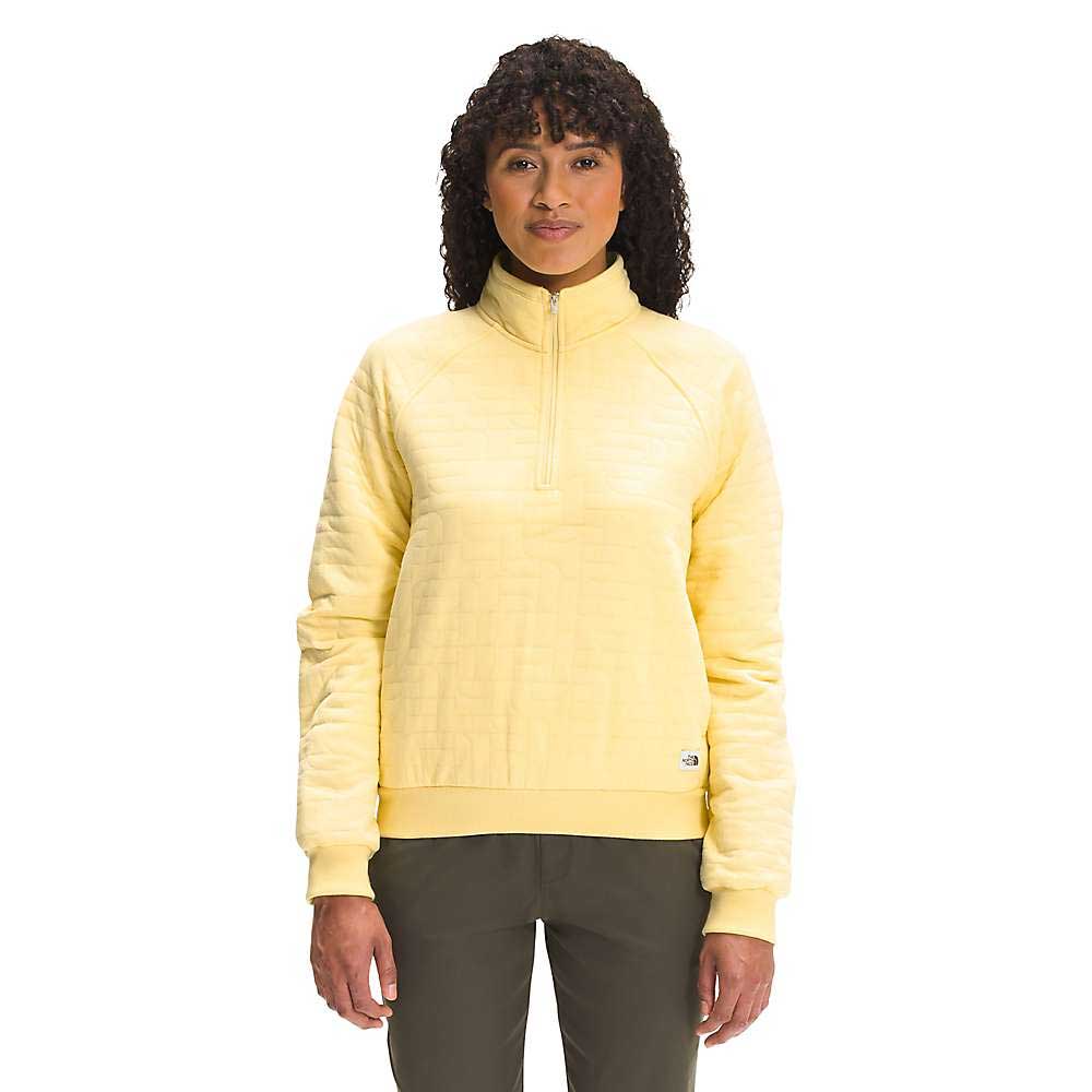 A woman with curly hair faces forward against a plain white background, wearing The North Face Women's Longs Peak Quilted 1/4 Zip Jacket in light yellow and olive green pants.