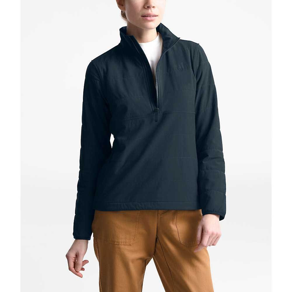 A person wears a black North Face Women's Mountain Sweatshirt 3.0 Pullover over a white shirt with tan pants, standing against a white background. Their head is slightly tilted, and their right hand rests relaxed by their side.