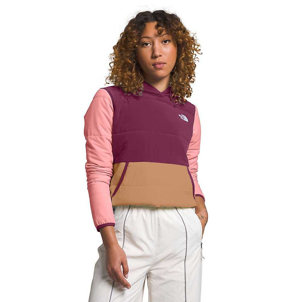 A woman with curly hair wears The North Face Womens Mountain Sweatshirt Pullover in color-blocked burgundy, pink, and tan with white pants, standing against a plain white background with one hand in her pocket.