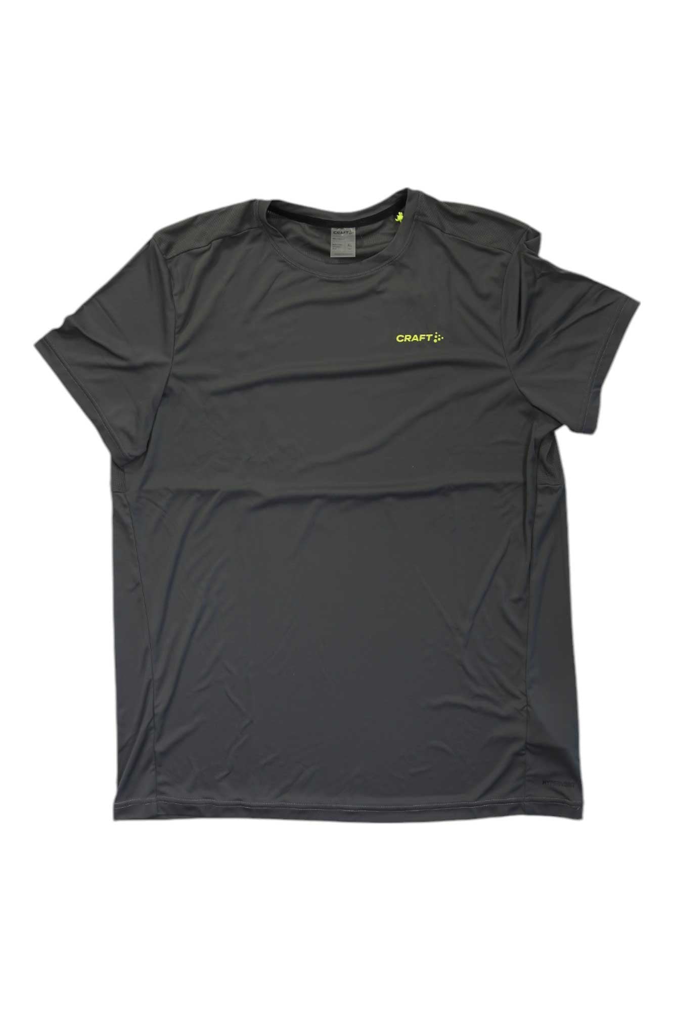 The Craft Sportswear Mens Core Essence SS Tee is a plain, short-sleeved, dark gray recycled jersey crew neck with a small yellow CRAFT logo on the left chest, designed for moisture-wicking comfort. Laid flat on a white background.