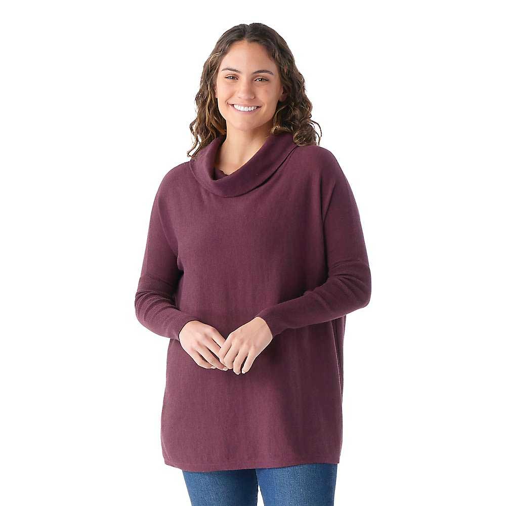 Smiling woman with long curly hair wears the Smartwool Womens Edgewood Poncho Sweater in plum and blue jeans, standing against a plain white background.