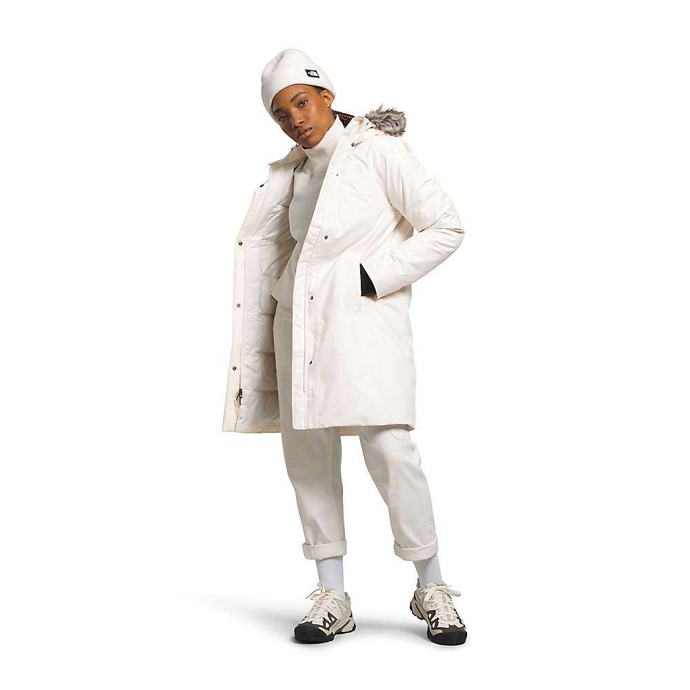 A person faces the camera wearing The North Face Womens Arctic Parka—a long white coat with a fur-trimmed hood—paired with a white beanie, turtleneck, pants, gray socks, and white sneakers with black soles.