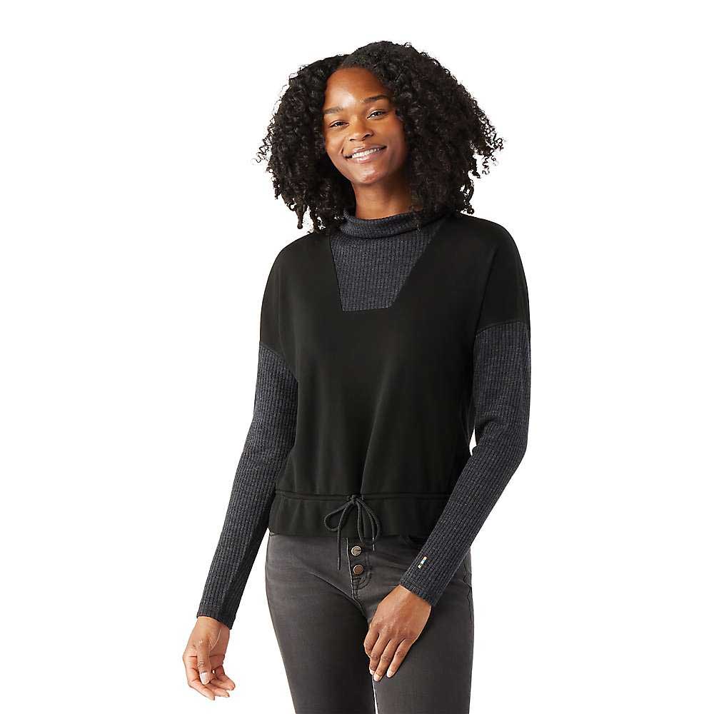 A woman with curly hair smiles in the Smartwool Womens Thermal Merino Rib Top—black, with ribbed sleeves and a drawstring waist—paired with dark jeans against a white background. Ideal for layering on snowsports adventures.