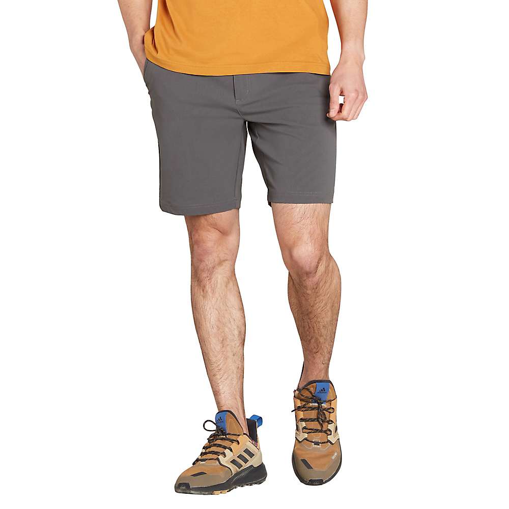 A person wears Sherpa Men's Bara Shorts in Kharani gray and brown hiking shoes with blue accents, standing against a white background. A mustard yellow shirt is partly visible, with only the lower body shown.