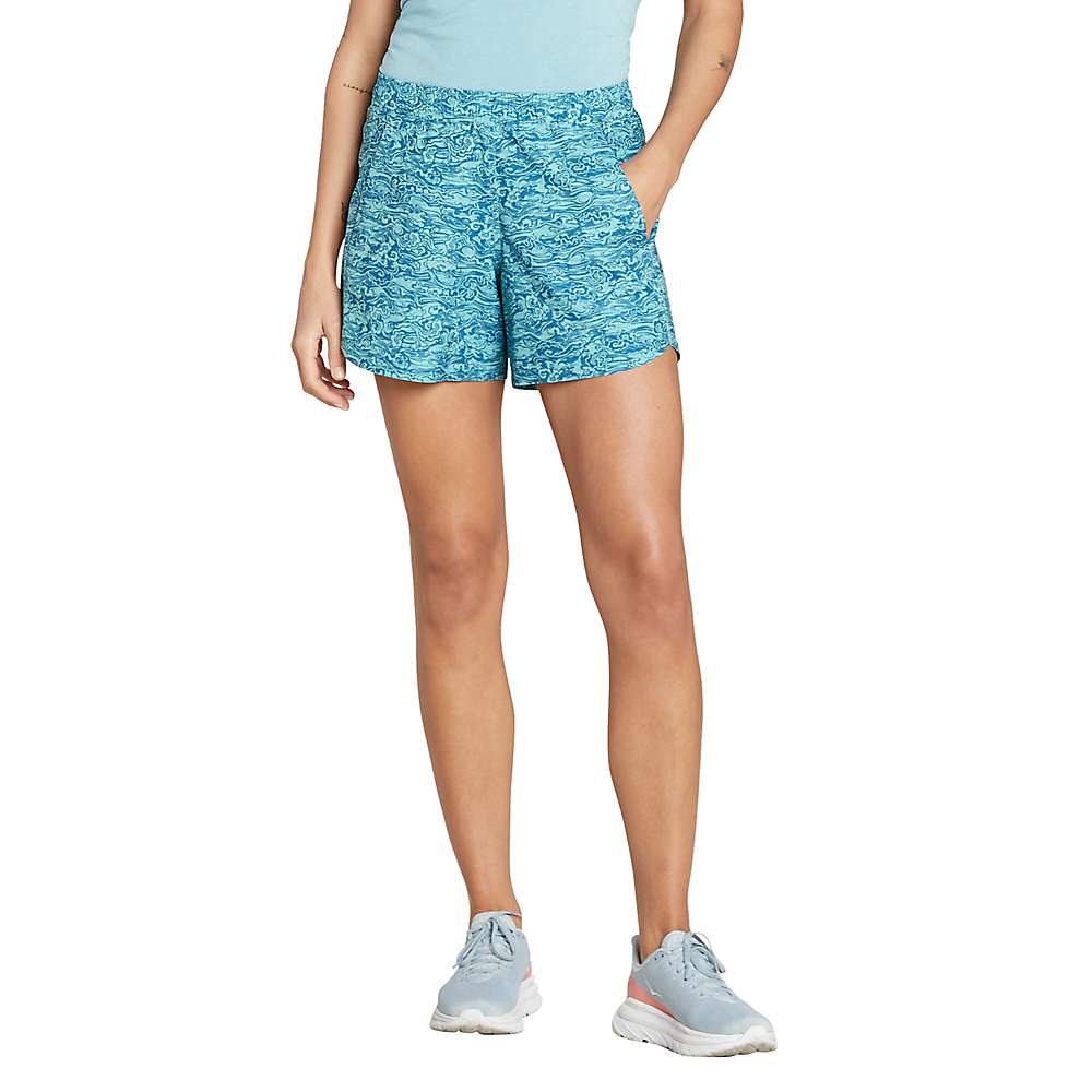 A person in a light blue shirt, Sherpa Women's Sajilo Pull On Shorts - Large Azule Cloud, and light gray sneakers stands with arms relaxed at their sides, facing forward against a white background.