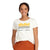 A woman with a side ponytail smiles while wearing the Topo Designs Women's Strata Map Tee - Large Natural, a white organic cotton shirt featuring yellow mountains, striped lines, and "Topo Designs Colorado, USA" on the front.
