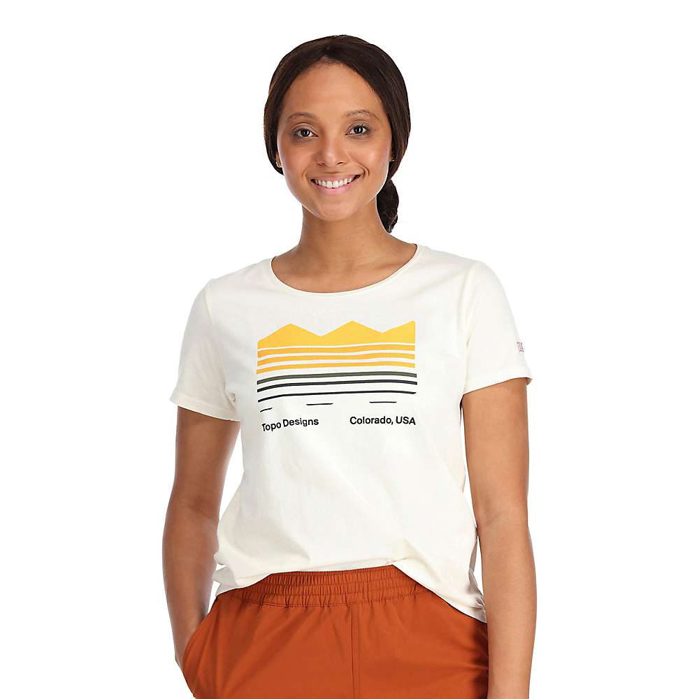 A smiling woman wears the Topo Designs Women's Strata Map Tee - Large Natural, an organic cotton t-shirt with a map graphic and "Topo Designs Colorado, USA," paired with rust-colored pants against a white background.