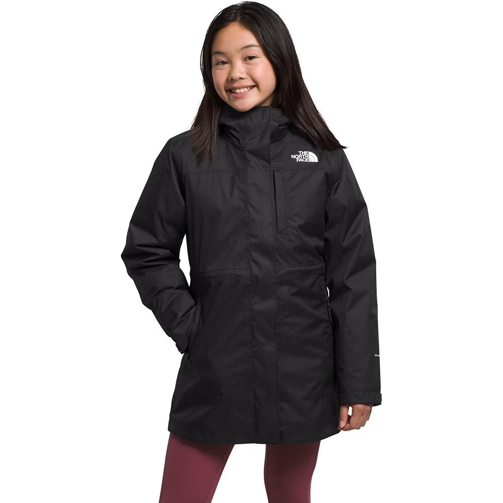 A young girl with long dark hair smiles in maroon leggings and The North Face Girls North Down Triclimate Jacket, a long black recycled outdoor coat with a high collar, her left hand in the pocket, against a plain white background.
