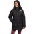 A young girl with long dark hair smiles in maroon leggings and The North Face Girls North Down Triclimate Jacket, a long black recycled outdoor coat with a high collar, her left hand in the pocket, against a plain white background.