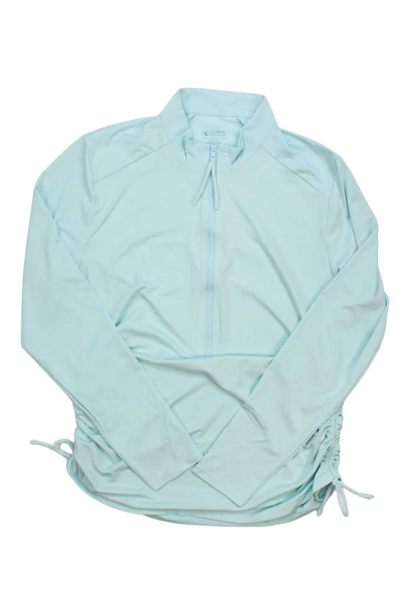 The Carve Designs Women's Cruz Rash Guard is a light blue long-sleeve pullover with a half-zip front and adjustable side drawstrings, offering UPF 50+ sun protection—ideal as a swim shirt for outdoor activities. Shown laid flat on white background.