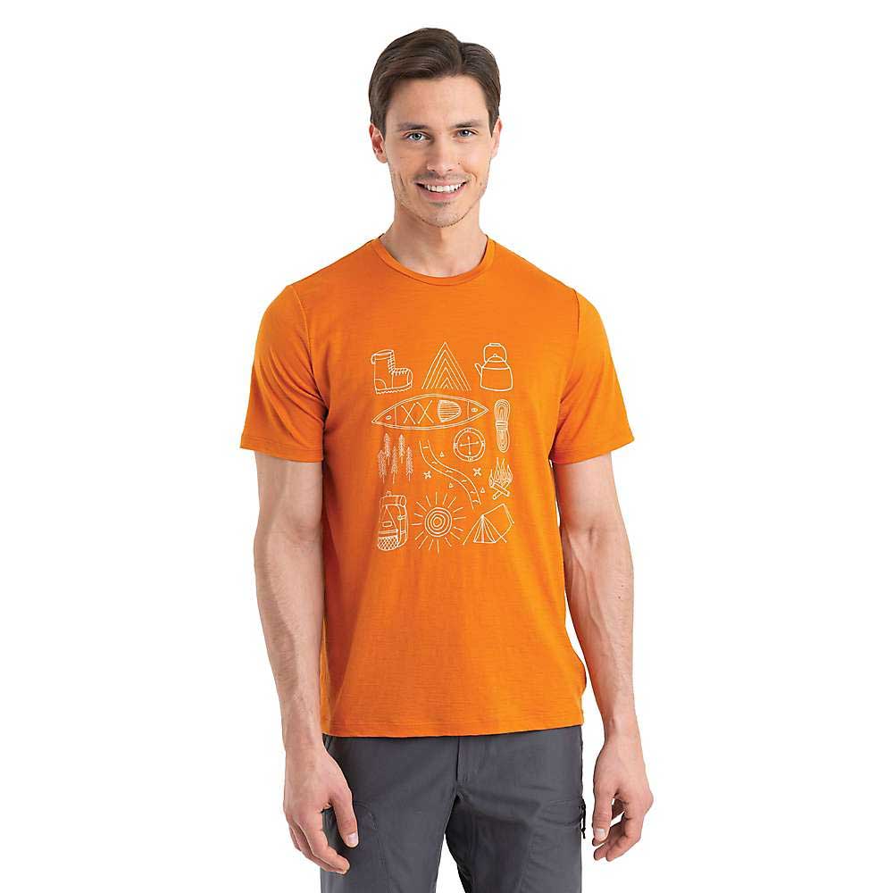 A man smiles wearing the Icebreaker Mens Merino 150 Tech Lite II Camp Essentials SS Tee in orange, featuring white outdoor-themed line drawings, along with gray pants and his arms relaxed at his sides.