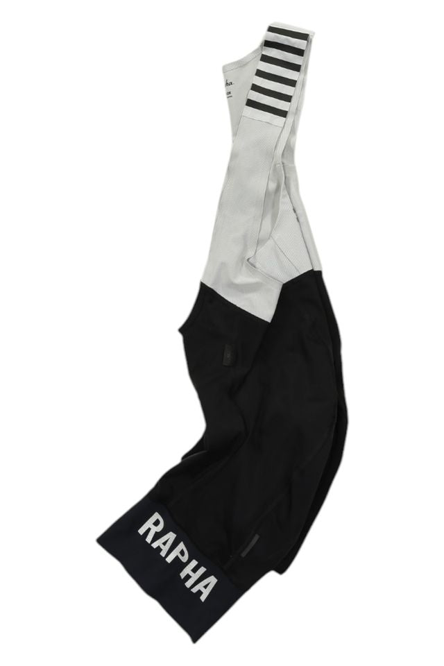 The Rapha Mens Pro Team Bib Shorts II - Regular are black and white with a large white RAPHA logo on the left leg, race-grade chamois, and a white mesh upper, shown laid flat on a white background.