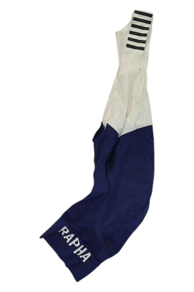 A pair of Rapha Mens Pro Team Bib Shorts II - Regular with a race-grade chamois, white upper, navy blue lower, striped strap, and RAPHA in white on the leg.