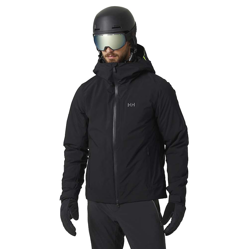 A person wearing a Helly Hansen Men's Swift Infinity Jacket, black gloves, black pants, a black helmet, and reflective ski goggles stands against a white background.