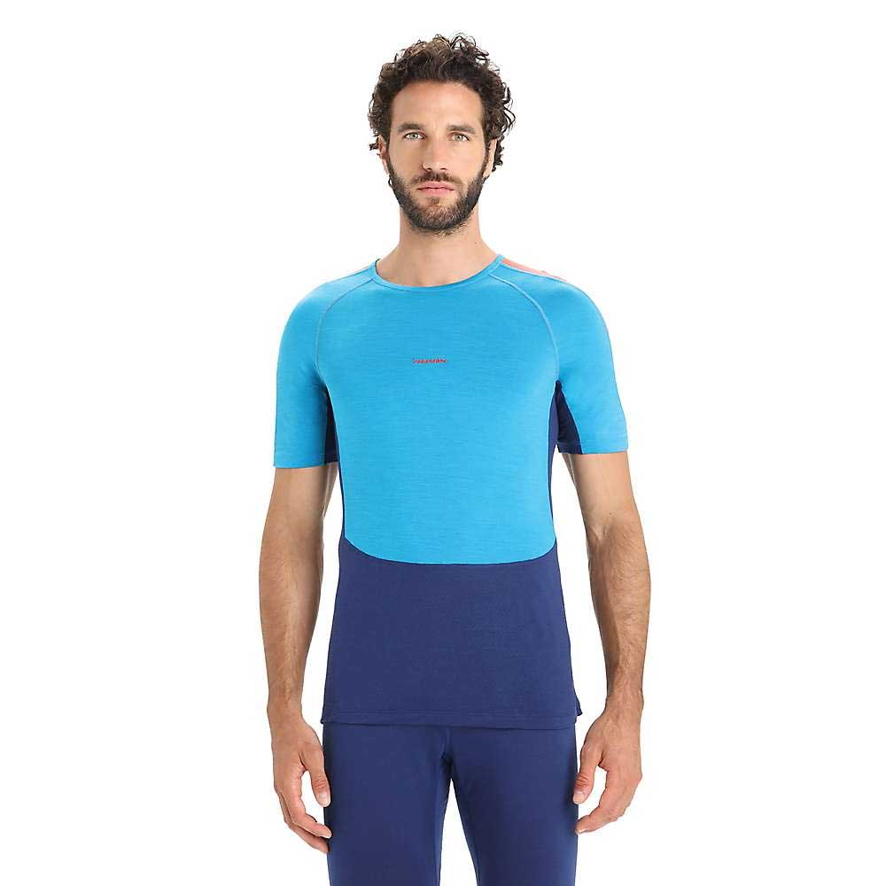 A man with short curly hair and a beard is wearing the Icebreaker Mens 125 ZoneKnit SS Crewe in two shades of blue with matching pants, standing against a white background.