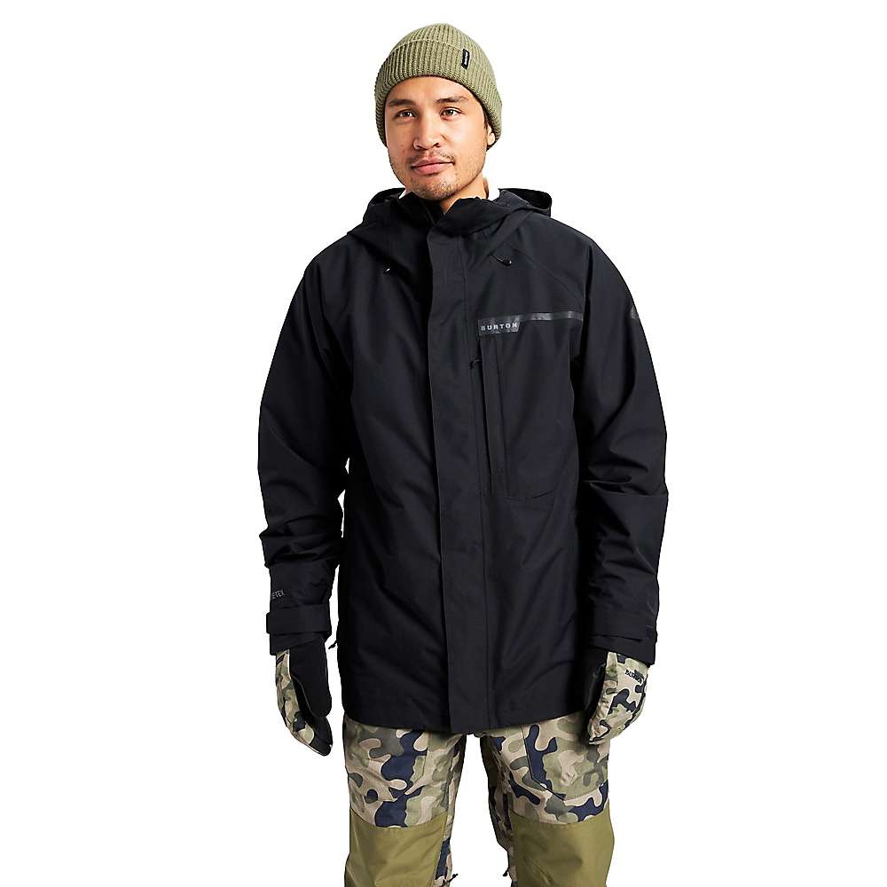 A person wearing a green knit beanie, a Burton Mens GTX Powline Jacket, camouflage gloves, and green camo-patterned pants stands against a plain white background.