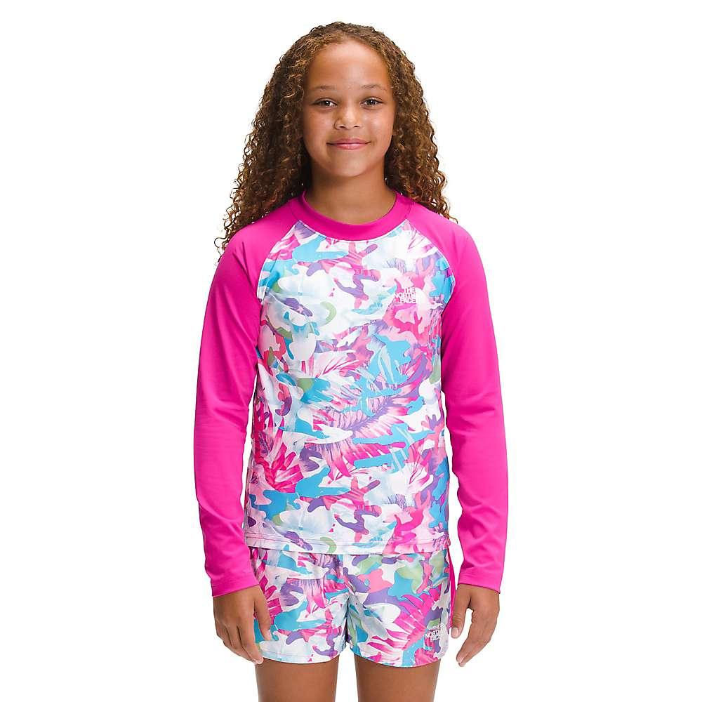 A young girl with curly hair smiles against a white background, wearing The North Face Girls Printed Amphibious LS Sun Tee—a pink and multicolored long-sleeve rash guard with UPF 50+ sun protection.