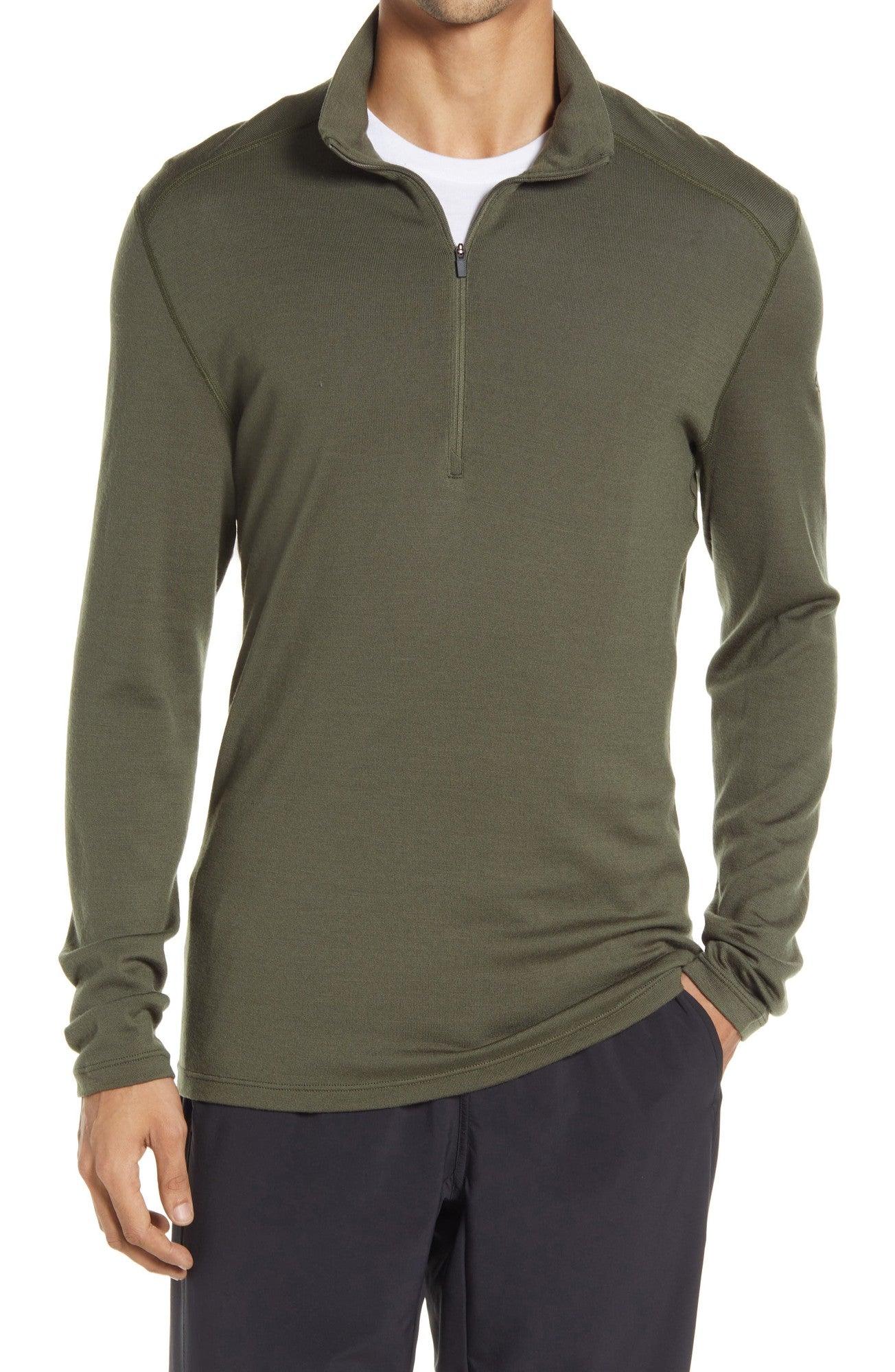 A man wearing the Icebreaker Mens 260 Tech LS Half Zip Top in olive green with a white undershirt and black pants, standing with one hand in his pocket.