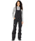 A woman with curly hair stands facing forward, wearing The North Face Womens Freedom Bib in black—featuring weatherproof fabric, adjustable straps, and a logo on one leg—with a light gray long-sleeve shirt underneath.