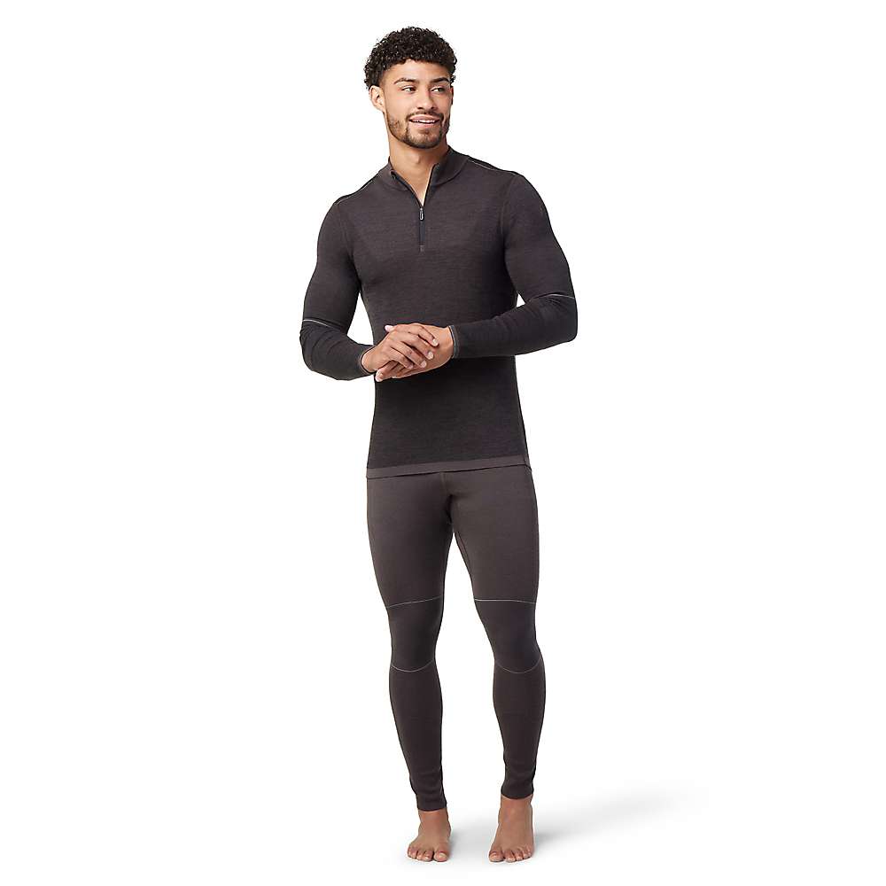 A man stands barefoot, slightly smiling against a white background, wearing the Smartwool Men's Intraknit Merino 250 Thermal 1/4 Zip top and matching fitted leggings as a dark merino wool base layer set.
