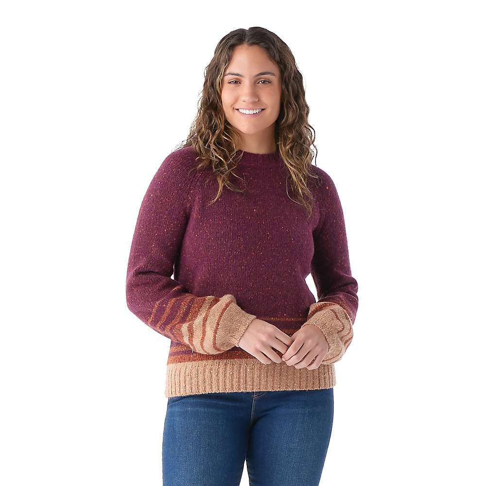 A woman with curly hair wears the Smartwool Women's Cozy Lodge Ombre Sweater in maroon with tan cuffs and waistband by Smartwool, standing smiling with hands clasped against a plain white background.