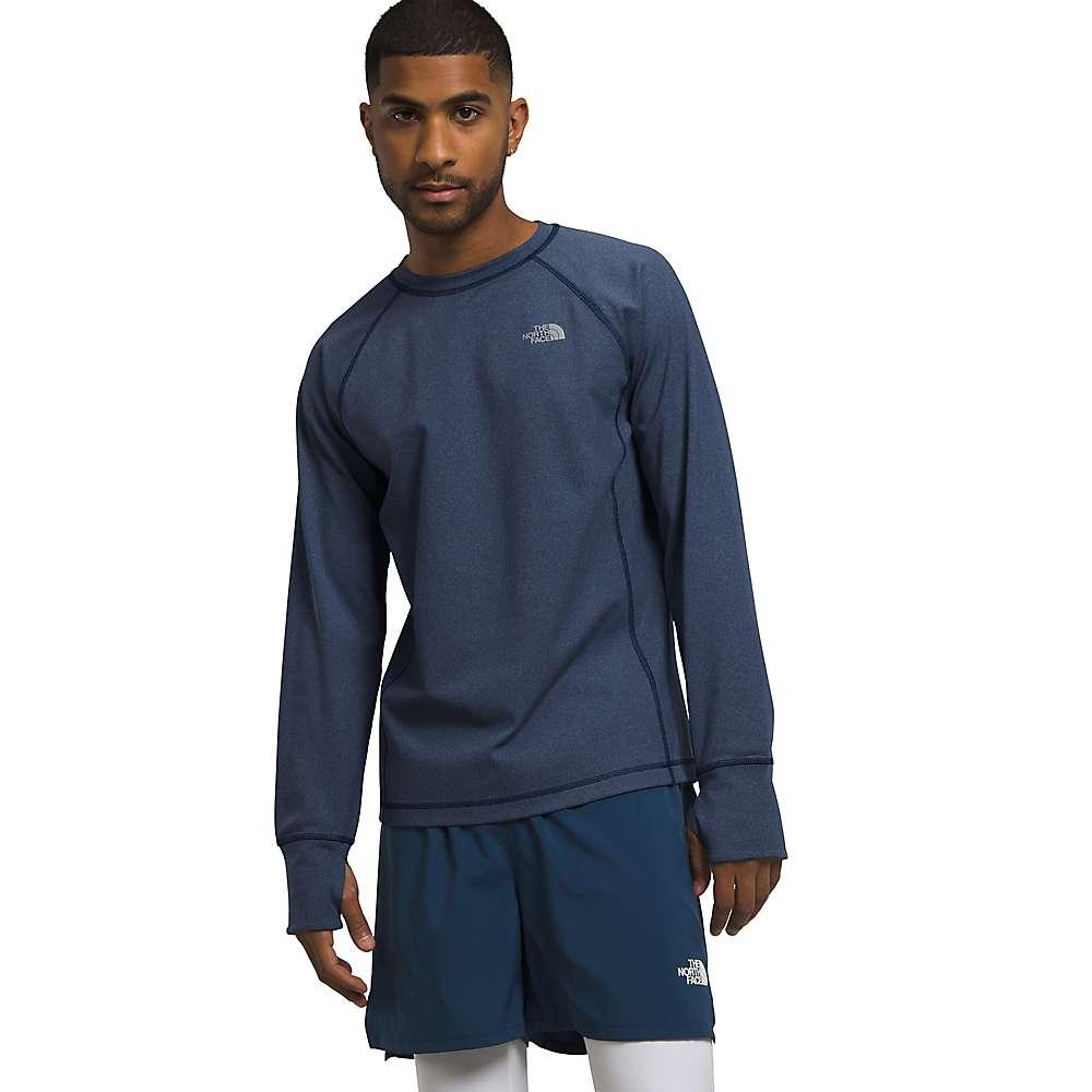 A man wears The North Face Men's Winter Warm Essential Crew in navy blue with moisture-wicking FlashDry-Pro technology and matching athletic shorts, standing against a white background and looking at the camera with one arm slightly bent.
