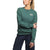 Wearing The North Face Womens Heritage Patch Crew in green, made from recycled fabric with a chest logo patch, and navy pants, a woman stands with one arm crossed, gazing to the side.