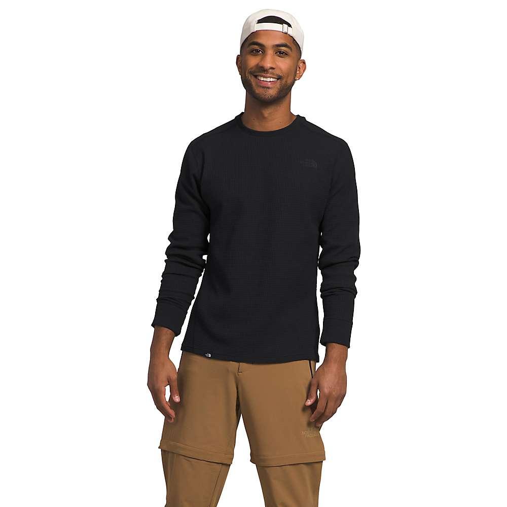 A man stands smiling against a plain white background, wearing The North Face Mens Canyon Fog Thermal LS Top in black, tan cargo pants, and a white backwards cap, with his right arm relaxed and his left hand in his pants pocket.