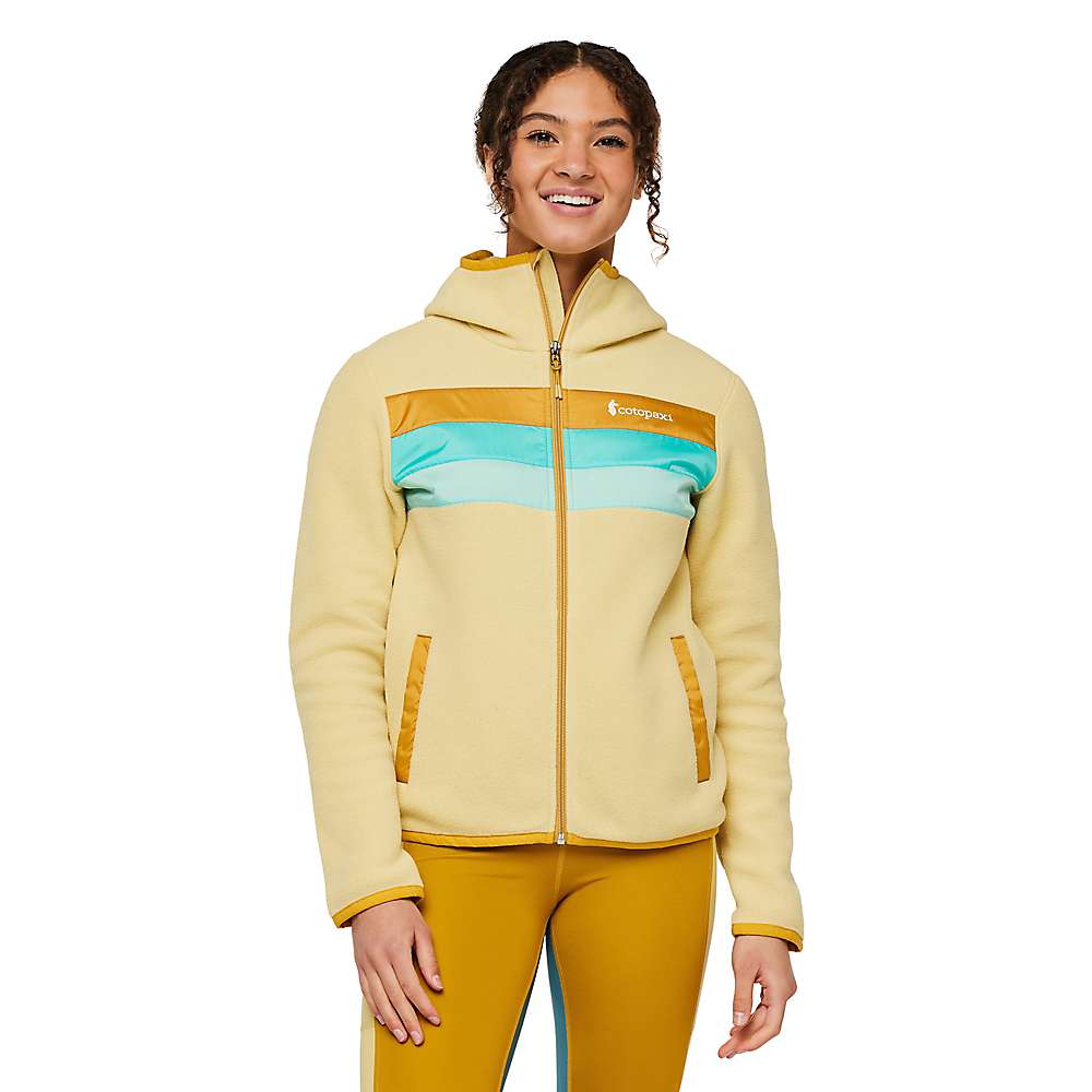 A woman smiles in a Cotopaxi Womens Teca Fleece Hooded Full-Zip Jacket, cream-colored with yellow, turquoise, and tan stripes and front pockets, paired with mustard yellow leggings against a white background.