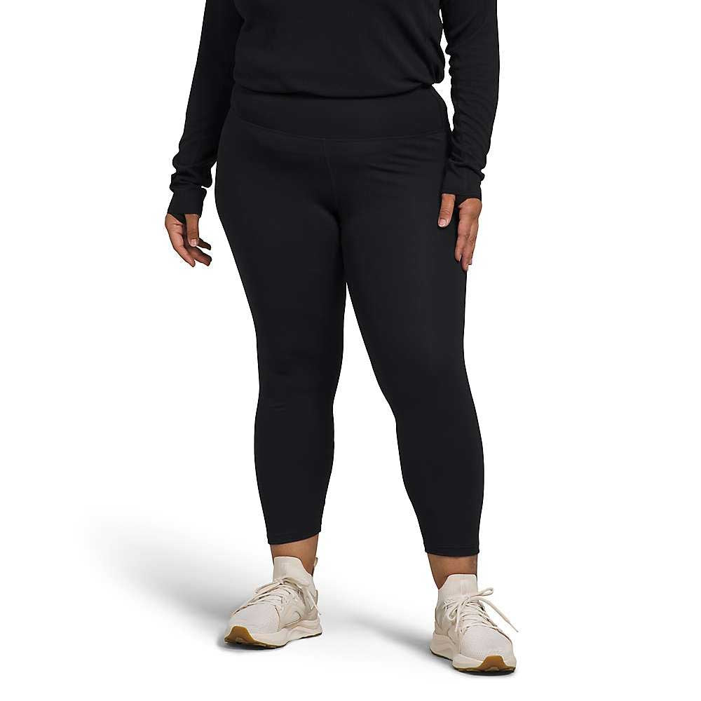 A person stands against a plain white background, wearing a black long-sleeve top, The North Face Womens Plus Fd Pro 160 Tight leggings made from recycled polyester, and cream sneakers. Their hands rest relaxed at their sides.