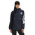 A woman stands against a plain white background wearing The North Face Womens IC Anorak Pullover, a relaxed fit dark hooded jacket with "USA" on the right sleeve and a stylized flag on the chest.