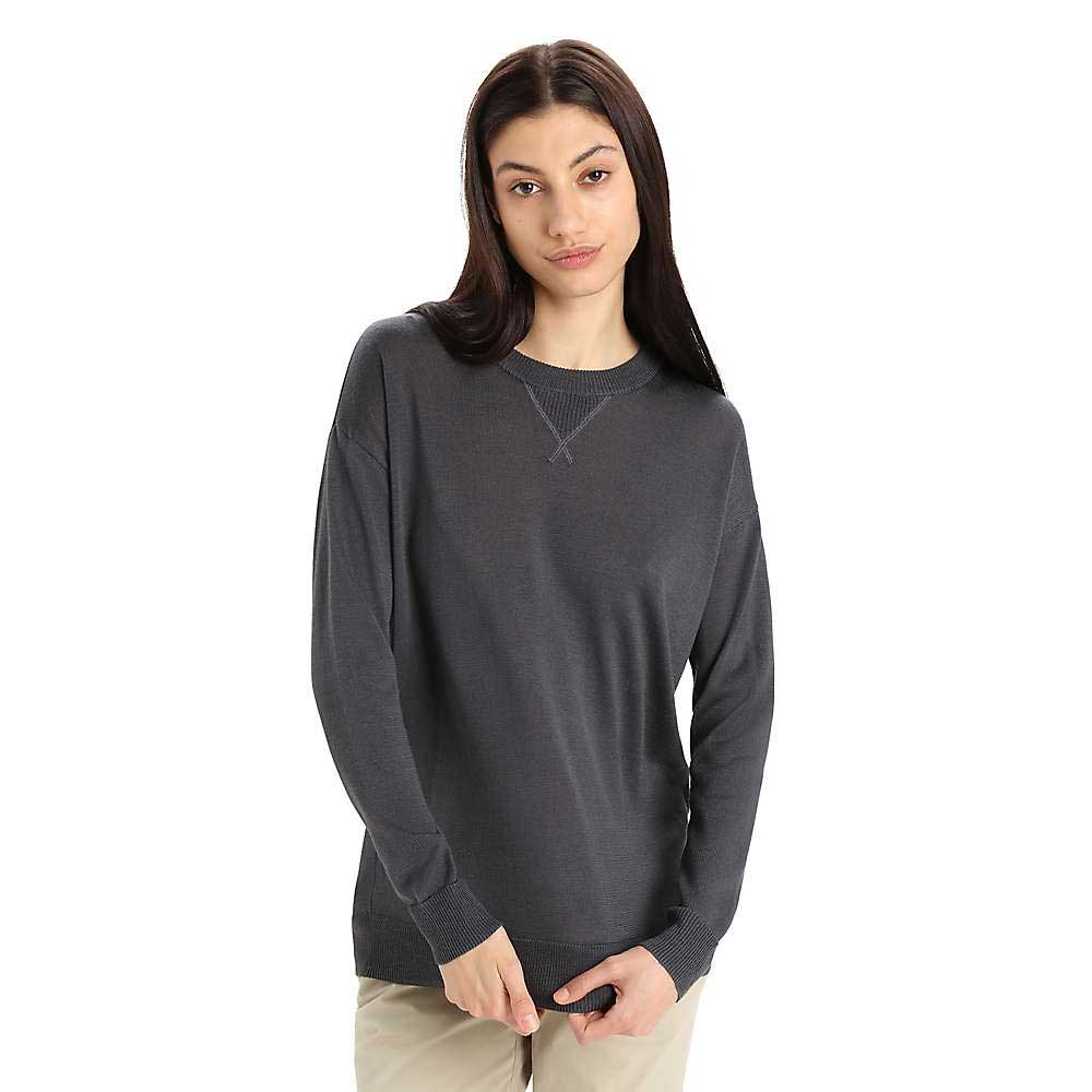 A woman with long dark hair and a neutral expression models the Icebreaker Womens Nova Sweater Sweatshirt in dark gray with light beige pants, standing against a plain white background, arms relaxed by her sides.