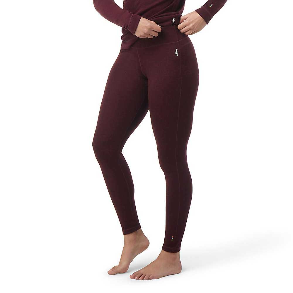 A person stands barefoot adjusting the waistband of Smartwool Womens Classic Thermal Merino Base Layer Bottom leggings in dark burgundy, paired with a matching long-sleeve top against a plain white background.