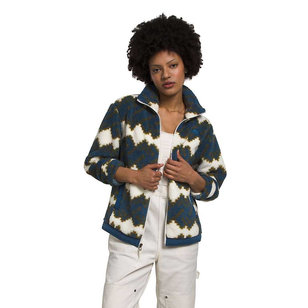 A woman with curly hair wears The North Face Womens Campshire Fleece Jacket, made from recycled polyester in blue, white, and olive-green patterns, over a white outfit against a plain white background.