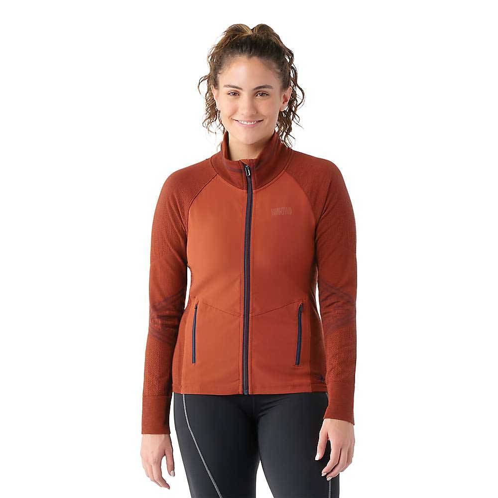 A woman with curly hair in a bun smiles while wearing the Smartwool Womens Intraknit Active Full Zip Jacket in rust and black leggings, standing against a plain white background.