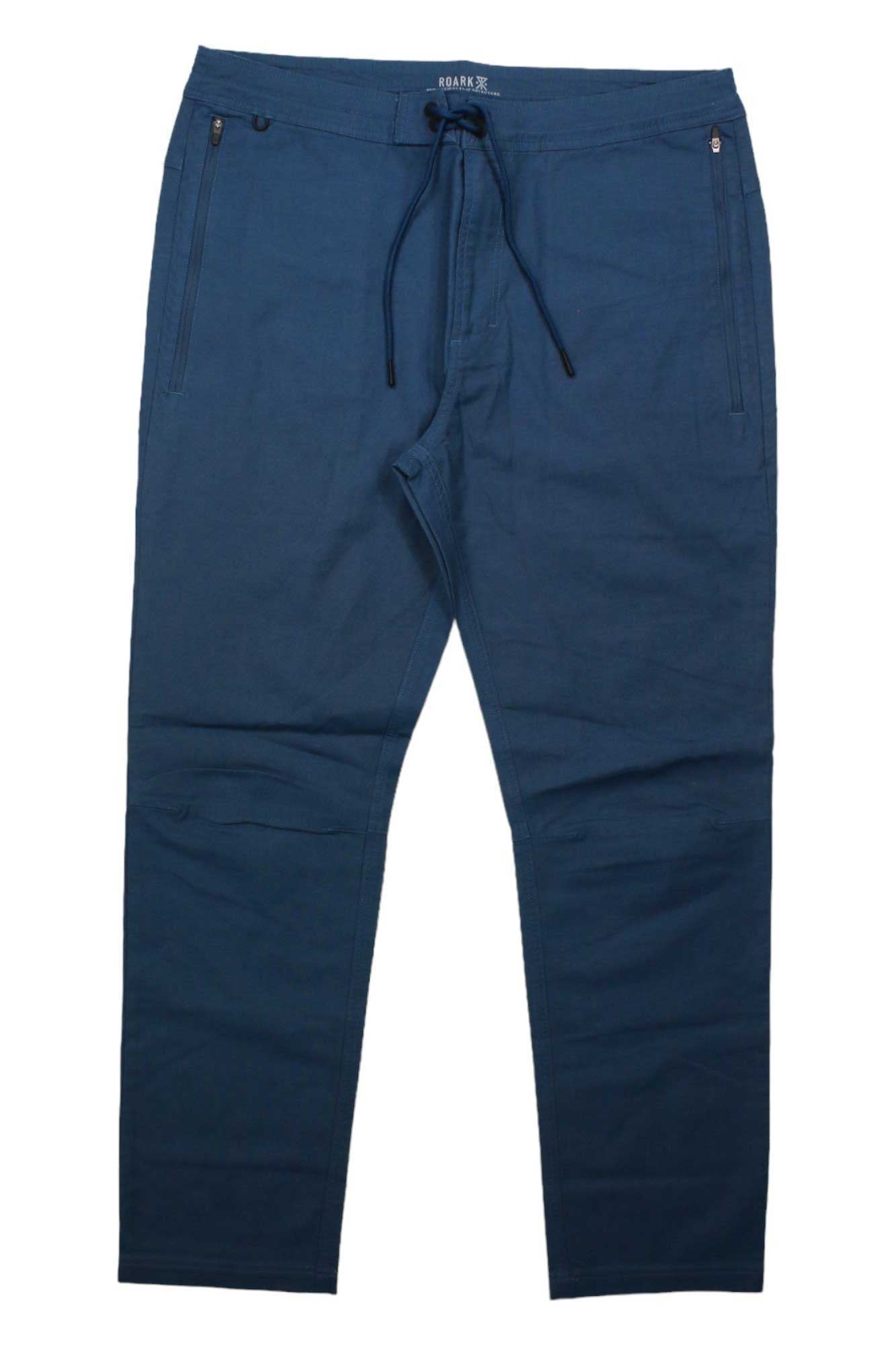 The Roark Men's The Layover 2.0 Pant in blue stretch canvas features zippered side pockets, a drawstring waist, straight legs, and a relaxed fit, shown laid flat on a white background.