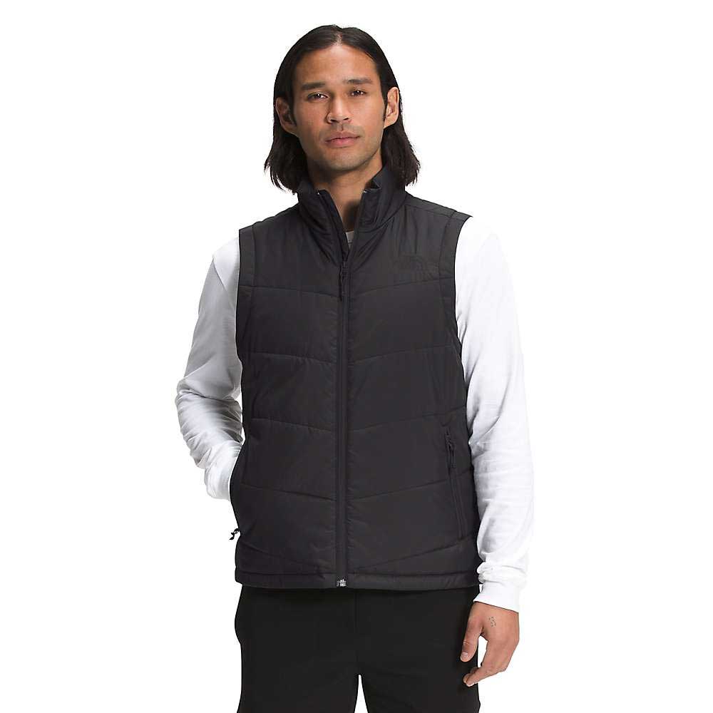 A person with long dark hair wears The North Face Men's Junction Insulated Vest in black over a white long-sleeve shirt and black pants, standing against a plain white background.
