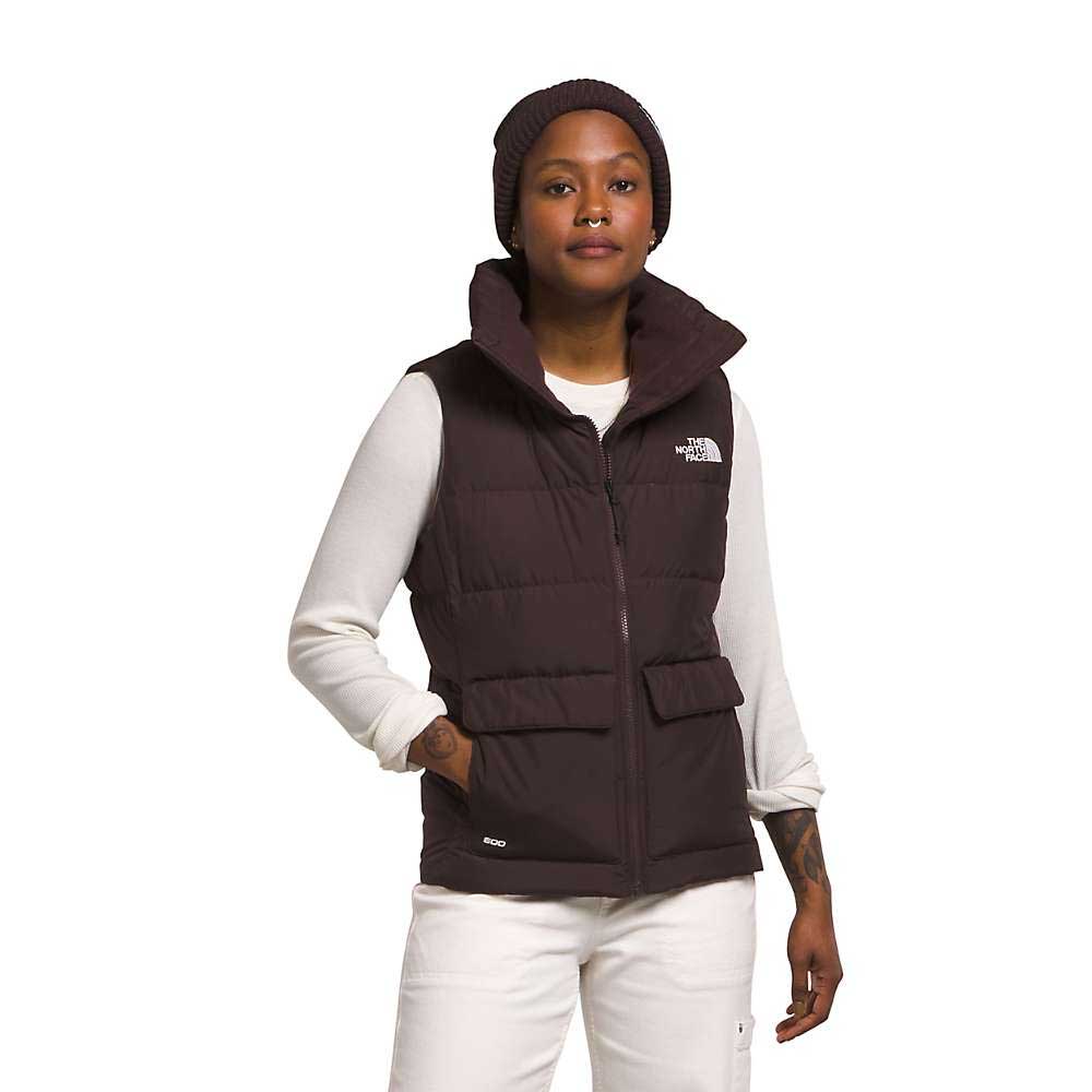 Wearing The North Face Womens Gotham Vest with recycled waterfowl down, a dark beanie, white pants, and a long-sleeve shirt, a woman stands against a white background and looks at the camera with a relaxed expression.
