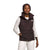 Wearing The North Face Womens Gotham Vest with recycled waterfowl down, a dark beanie, white pants, and a long-sleeve shirt, a woman stands against a white background and looks at the camera with a relaxed expression.