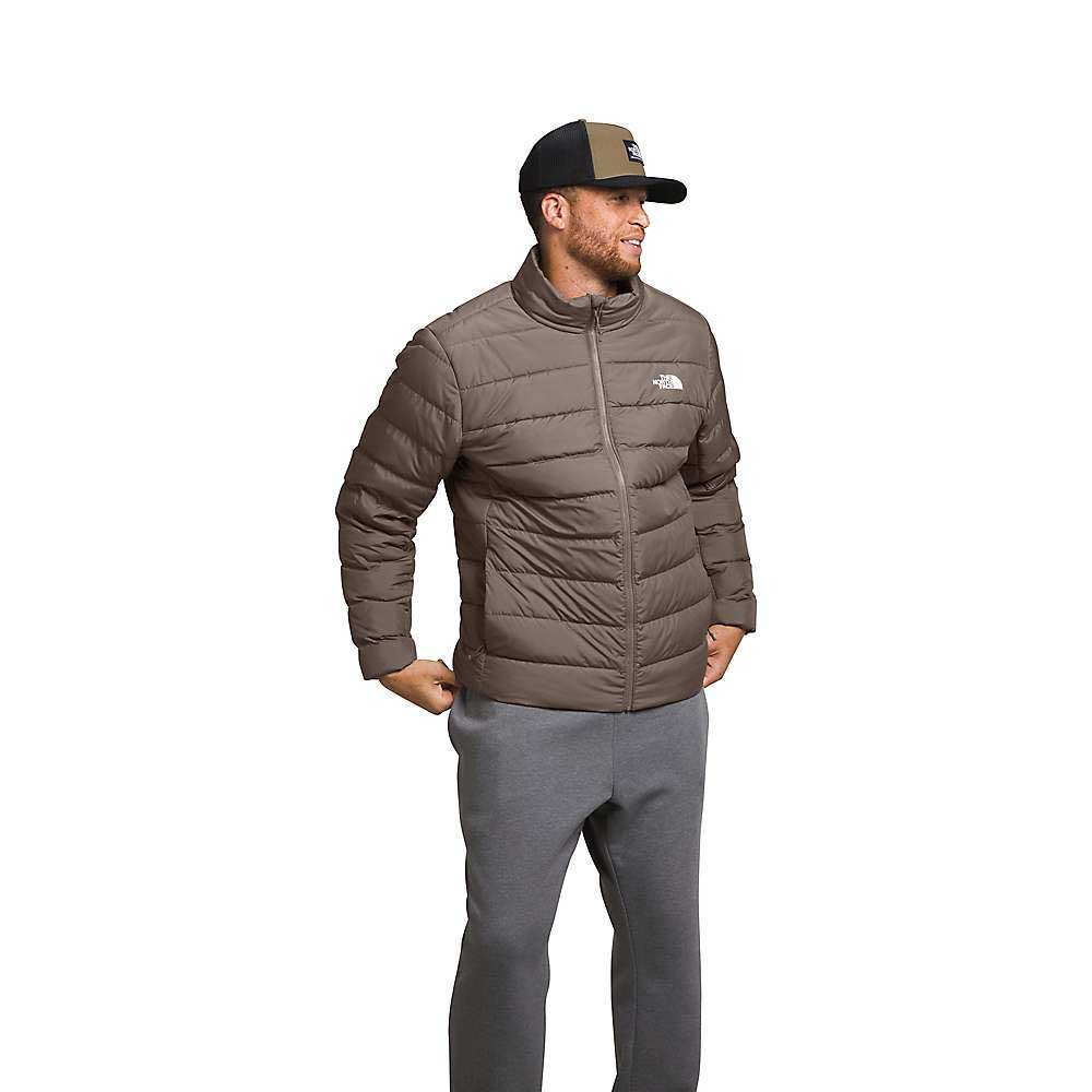A man wearing The North Face Men's Big Aconcagua 3 Jacket in brown, gray sweatpants, and a black and beige baseball cap stands smiling with his hands in his pockets against a white background.