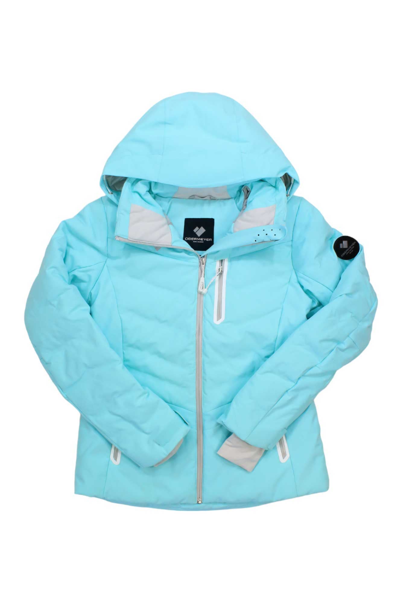 The Obermeyer Womens Cosima Down Jacket is a light blue insulated winter coat with a hood, front zipper, zippered side and chest pockets, a black patch on the left sleeve, and a quilted design for added warmth.