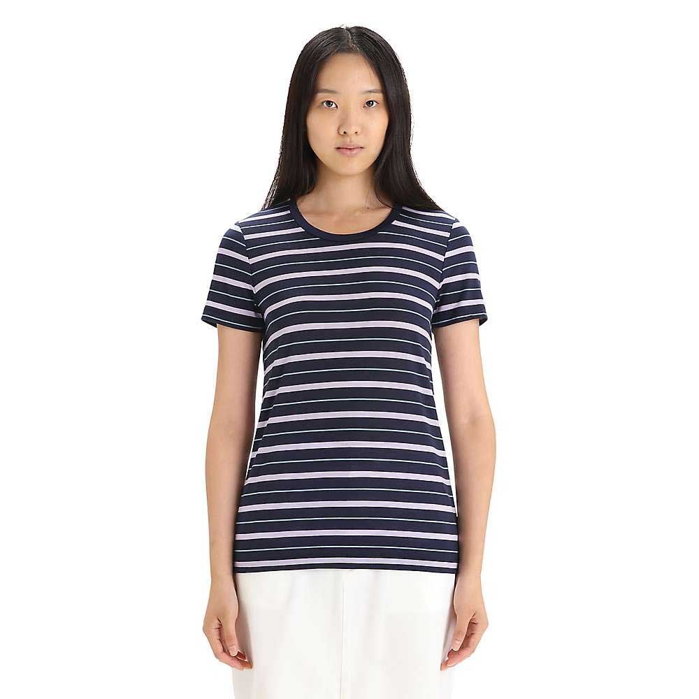 A person with long black hair wears the Icebreaker Womens Wave SS Stripe Tee, a navy blue top with white and light pink stripes made from Cool-Lite Merino Wool, paired with white pants and standing against a plain white background.