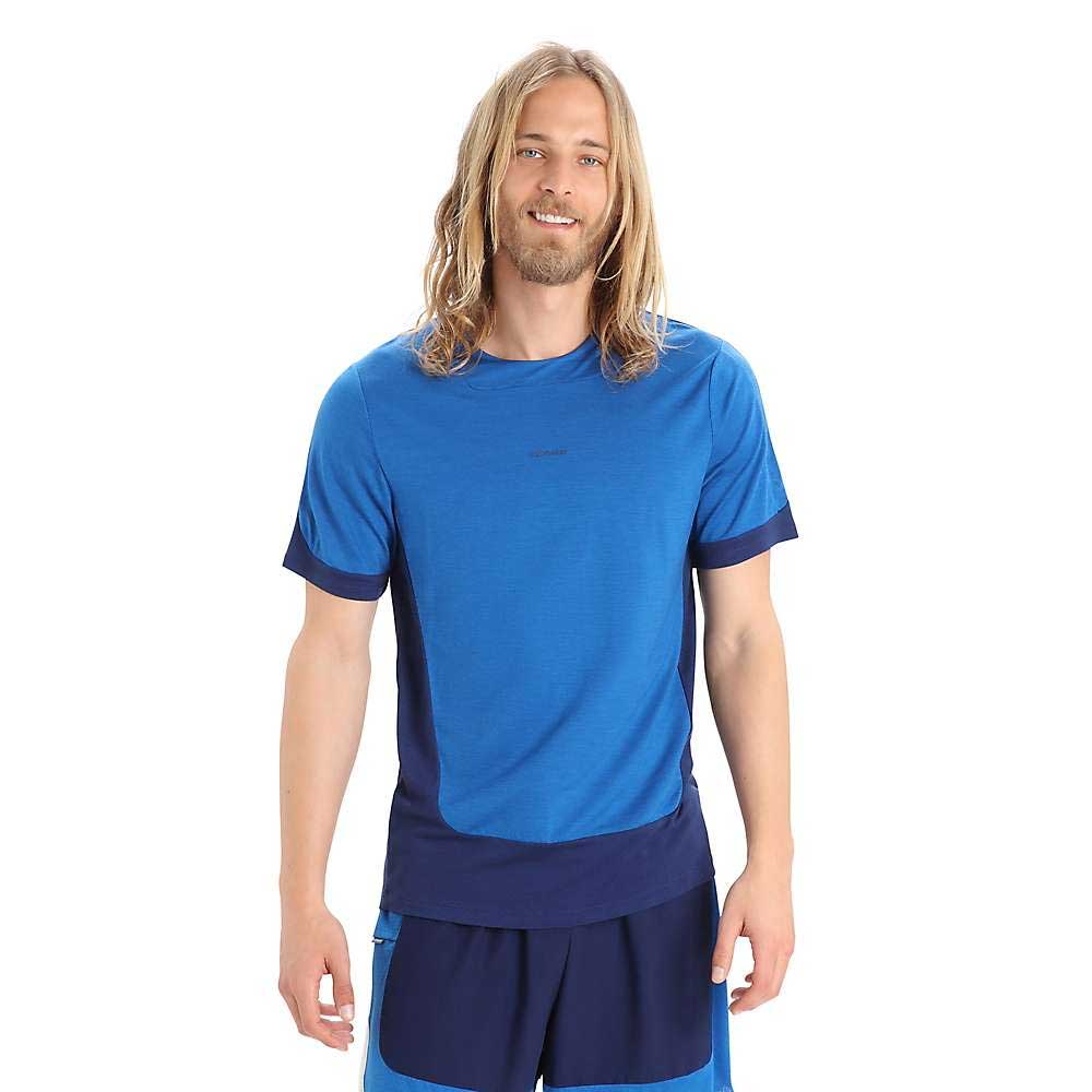 A man with long blonde hair and a beard is smiling against a white background. He is wearing the Icebreaker Mens Zoneknit SS Tee, a blue breathable T-shirt with dark blue accents, paired with matching blue shorts.
